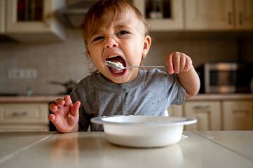 Baby boy eating porridge with spoon in kitchen