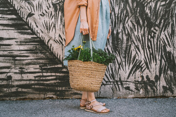 Woman with leafy vegetables and flowers in wicker bag by patterned wall