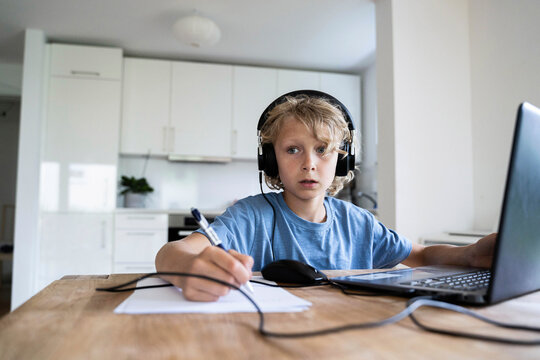 Thoughtful boy with wired headphones studying through laptop at home