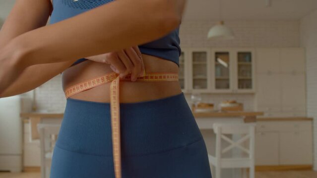 Midsection of slimming African American woman in sportswear measuring waist with tape, checking progress of dieting plan while standing in domestic kitchen. Close-up.