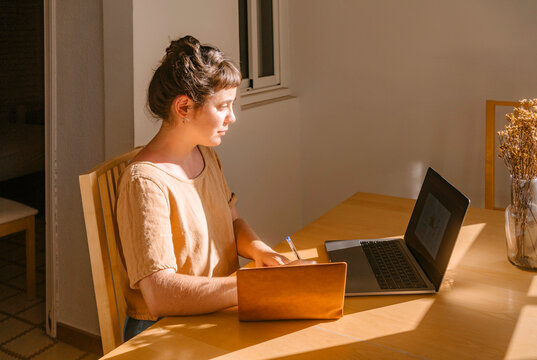 Graphic designer writing in book with laptop at desk - Powered by Adobe