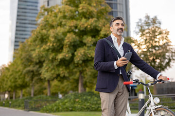 Smiling businessman with smart phone holding bicycle on footpath