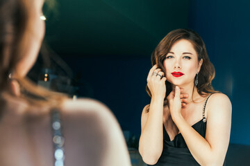 Portrait of posh woman with makeup and stylish dark hair in elegant cocktail silk black dress looking on herself in mirror reflection. Fashionable and self-confident lady. Self Assurance