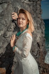 Middle aged woman looks good with blond hair, boho style in white long dress on the beach decorations on her neck and arms.