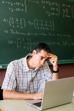 Stressed math teacher sitting at table with laptop in classroom