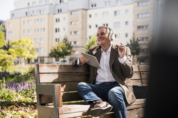 Senior man with tablet PC enjoying music listening through wireless headphones on bench