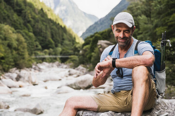Smiling man checking time on wristwatch sitting in front of mountains