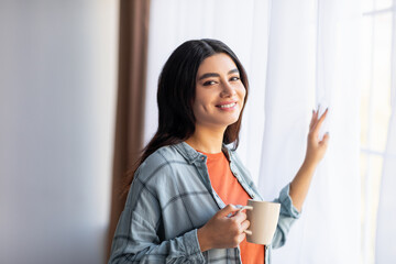 Happy young Arab lady drinking fresh coffee near window at home, enjoying relaxing morning