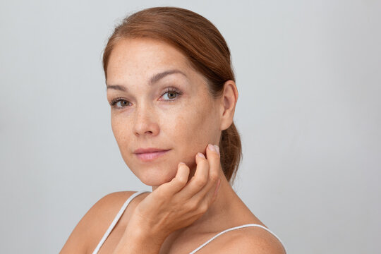 Portrait of cropped caucasian middle aged woman face with freckles holding fingers on cheek, showing soft skin on white background looking at camera