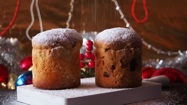 Sprinkling powdered sugar over traditional Christmas mini Panettone with raisins and dried fruits on golden background  surrounded by red baubles and garlands.