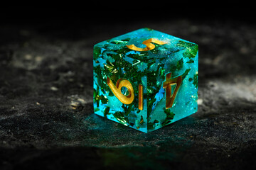 A turquoise dice with gold numbers for a board game on a dark stone top
