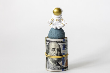 A rolled-up roll of dollars and a figure of an astronaut meditating on the planet. White...