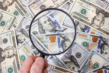 A hand with a magnifying glass against the background of scattered American dollar bills, close-up....