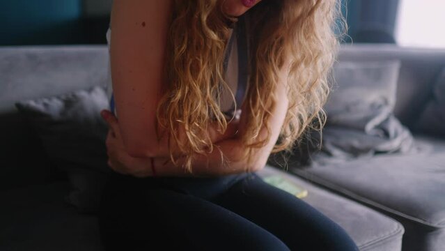 Woman suffering from severe stomach ache sitting on sofa in living room, feeling extremely sick. Young adult female experiencing period cramps. Girl suffering from sharp bladder pain. Close up.