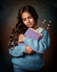A girl in a blue sweater holds old books in her hands. The book includes a bookmark of dried...