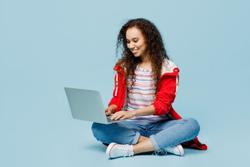 Full body young IT woman of African American ethnicity wear red jacket sitting hold use work on laptop pc computer isolated on plain pastel light blue cyan background. Wet fall weather season concept.