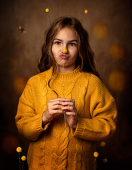 A girl in a yellow sweater holds a Kraspedia drumstick in her hands. She has horns on her head, a mustache made of flowers. A bee flies nearby. A kiss from the lips, a grimace. The girl has brown wavy