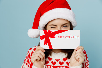 Young merry woman wear red sweater Santa hat posing cover mouth gift certificate coupon voucher card for store isolated on plain pastel light blue cyan background. Happy New Year 2023 holiday concept. - obrazy, fototapety, plakaty