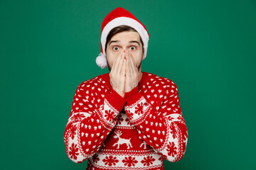 Merry young man 20s wear red warm knitted sweater Santa hat posing cover mouth with hand look camera isolated on plain dark green background studio portrait. New Year 2023 holiday celebration concept.