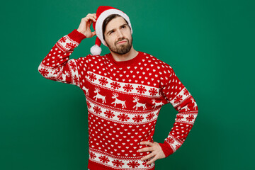 Merry puzzled thoughtful young man wear red warm knitted sweater Santa hat posing look aside hold scratch head isolated on plain dark green background studio New Year 2023 holiday celebration concept