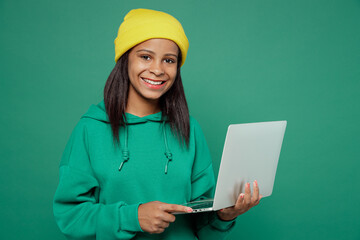 Little smiling happy IT kid teen girl of African American ethnicity 13-14 years old wear casual hoody hat hold use work on laptop pc computer isolated on plain dark green background Childhood concept.