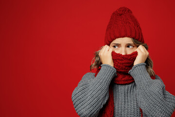 Young sad woman wear warm gray sweater cover mouth with scarf hat look aside on area isolated on plain red background studio portrait. Healthy lifestyle ill sick disease treatment cold season concept.