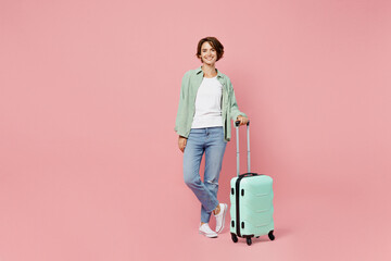 Full size young traveler woman wear green shirt hold suitcase isolated on plain pastel light pink color background Tourist travel abroad in free spare time rest getaway Air flight trip journey concept