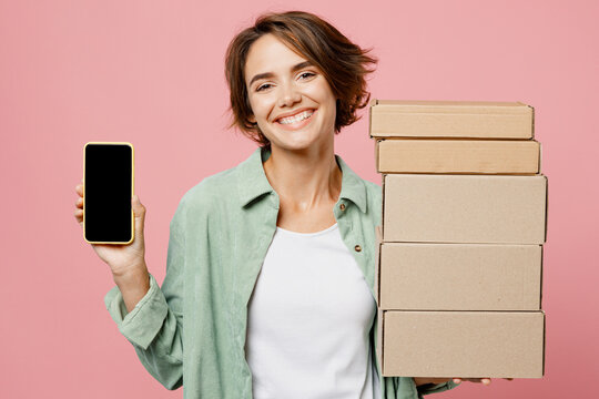 Young woman wear green shirt white t-shirt hold in hand use mobile cell phone with black screen area brown clear blank craft paper cardboard box mock up isolated on plain pastel light pink background
