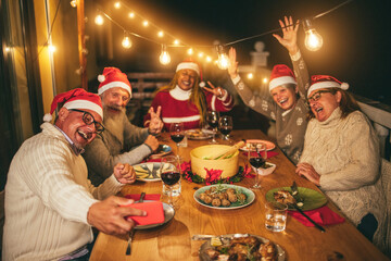 Happy senior friends taking a selfie during Christmas dinner at home wearing Santa Clause hats -...