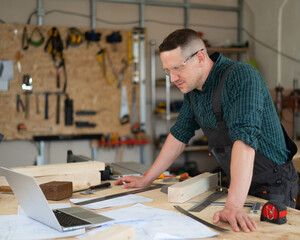 Portrait of a carpenter in protective glasses and work overalls uses a laptop in a workshop.