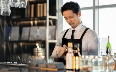 Asian bartender man standing at bar counter prepare to service cocktail alcohol at restaurant, portrait. Handsome young barman in uniform working at nightclub. Waiter person in nightlife lifestyle.
