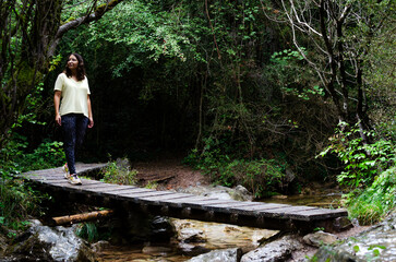 young woman with glasses crossing the river on a bridge between trees and rocks. strong, free and self-confident, she is hiking alone.