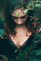 Brunette woman with head looking down, eyes closed, wearing low cut black goth dress, surrounded with green ivy leaves