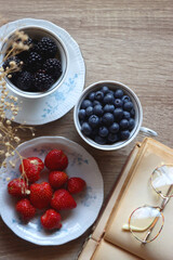 Vintage tea set filled with blueberries, blackberries and starwberries, open book, reading glasses and decorative flowers on the table. Flat lay.