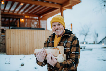 Senior man carrying wooden logs for preparing outdoor fire, during winter day.