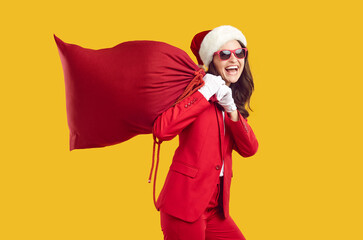 Funny modern Santa woman laughing loudly carrying big red bag with gifts on her shoulders. Woman in red suit, santa hat and sunglasses on orange background. Christmas shopping and sale concept.