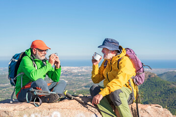 Happy elderly hikers with backpacks enjoying a cup of coffee taking a break, relaxing at the top of the hill