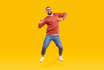 Fototapeta na wymiar Happy male dancer or fashion model in casual outfit dancing on yellow studio background. Funny cheerful young man in orange sweatshirt, blue jeans and glasses dancing and having fun
