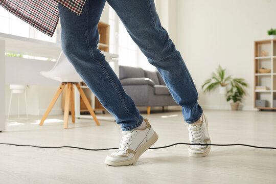 Legs of young man who, walking clumsily, stumbled over electric cord in office or at home. Man in sneakers and jeans trips over black power cord lying on floor. Low section.
