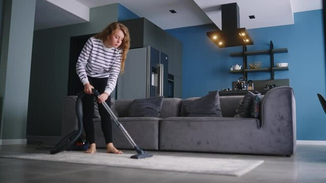 Young curly caucasian woman vacuums floor in her apartment. Adult female vacuum-cleaning the rug in modern stylish apartment. Process of cleaning the house. Housewife cleaned up the mess in the room.