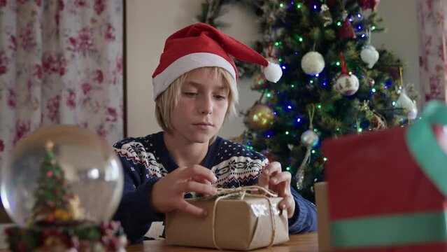 Kid with Santa hat wrapping Christmas gifts against Christmas tree.