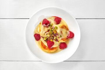 Sweet curd cheese pancakes with coconut custard, raspberries and pistachios on white wooden surface