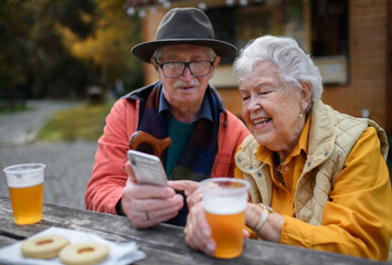 Happy senior couple in forest buffet resting after walk, having beer and looking at smartphone.