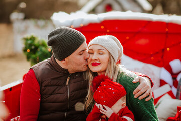 Happy family with dad, mom and newborn baby boy in winter clothes sitting in red Christmas car. Holiday concept.