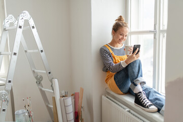 Happy young woman having break during renovation her aparatment. Using phone, searching inspiration...