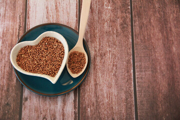 Obraz na płótnie Canvas Small grains of natural buckwheat in a decorative plate in the form of a heart and in a wooden spoon