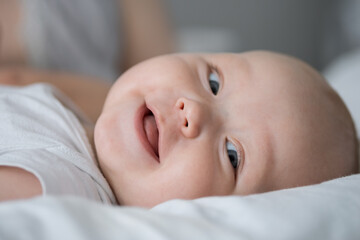 Baby girl with blue eyes looks around with smiling and happy expression lying on bed closeup. Excited baby enjoys parents care and laughs