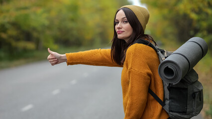 Woman hitchhiker with smiling expression waits for car and shows thumb at autumn forest. Brunette lady with backpack travelling by hitchhiking stands on road