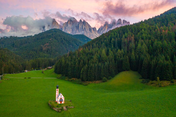 Fototapeta na wymiar St. Johann old church in the village Santa Magdalene against beautiful massif of the Dolomites, Odle or Geisler group in valley Val di Funes, covered with amazing pink clouds at sunset, aerial view