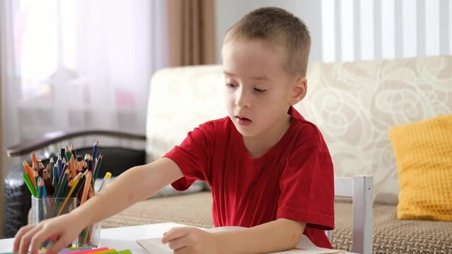 Portrait of funny happy little caucasian boy sitting at table and drawing with colored pencils and markers. Art therapy, rehabilitation, leisure.